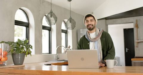 Indian man working on laptop in sunlit modern kitchen with arched windows and coffee mug