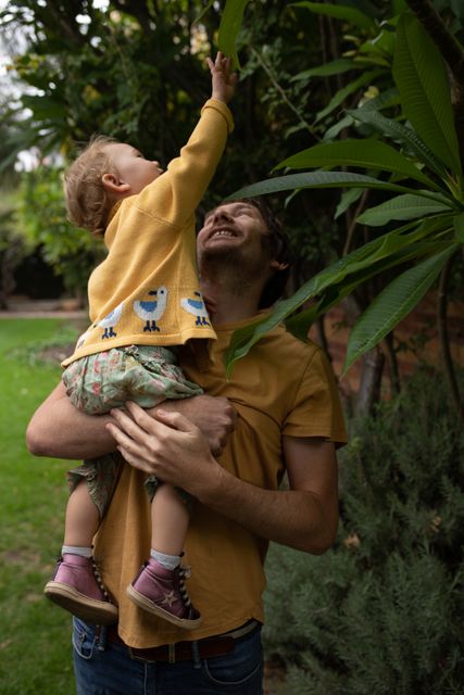 Front view of a young Caucasian father wearing yellow shirt, holding his baby, standing in the garden, baby touching the leaves, exploring.