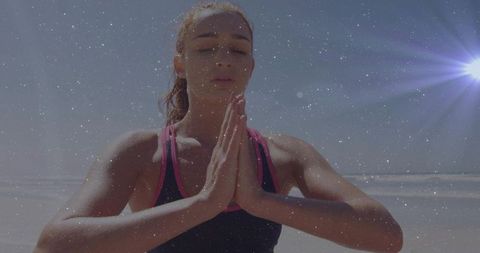 Meditating athletic woman finding calm on sunlit beach with prayer hands and sun flare