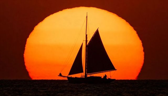 Single-mast sailboat silhouette sailing across sea at giant orange sun on horizon