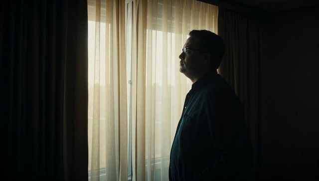 Contemplative man standing by sheer curtain at hotel window, backlit silhouette, moody interior