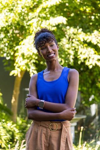 Smiling african woman standing with arms crossed in sunlit backyard garden wearing blue top