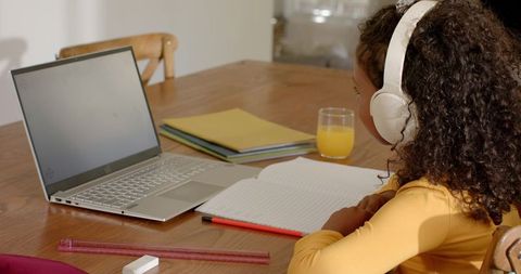 Young girl studying at home with laptop and headphones on wooden table, notebook open