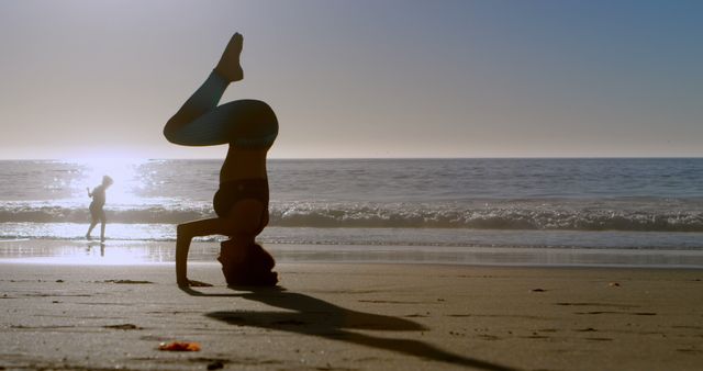Person practicing yoga headstand on beach at sunset, silhouette against the ocean horizon. Perfect for use in health and wellness blogs, meditation guides, fitness brochures, and lifestyle websites to showcase tranquility and healthy living.