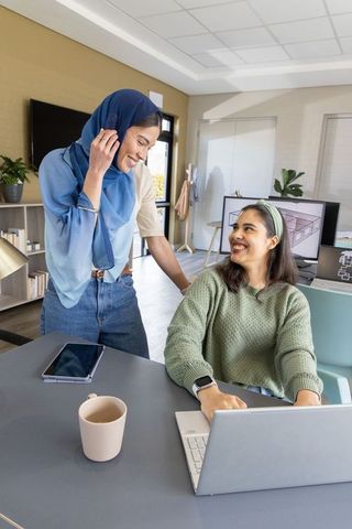 Collaborating female colleagues with hijab at modern office desk with laptop and monitors