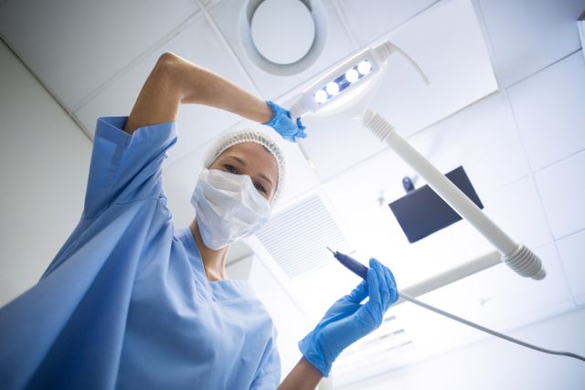 Dental assistant in surgical mask holding dental tools under dental light in a clinic. Ideal for use in healthcare, dentistry, and medical professional contexts. Can be used in articles about dental care, hygiene practices, and the role of dental assistants.