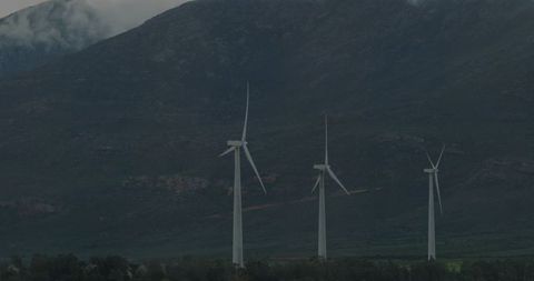 Wind turbines generating clean energy at misty mountain base in overcast rural landscape