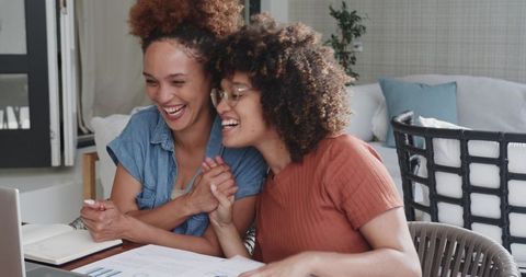 Laughing partners collaborating over laptop and printed charts at cozy home workspace