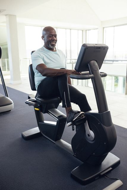 African American man exercising on a stationary bike in a bright, modern home gym. He is smiling and appears to be enjoying his workout. This image can be used for promoting fitness, healthy lifestyles, home gym setups, and wellness programs, especially targeting senior or older adults.