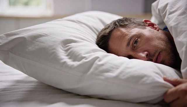 Adult man lying awake in bed holding pillow under duvet, contemplative morning light