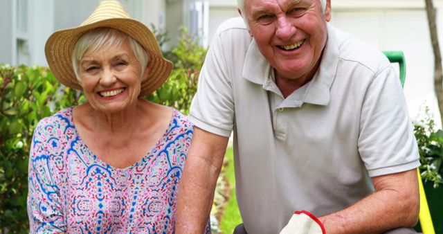This image features a joyous senior couple gardening outdoors on a sunny day. They appear very happy and engaged with their gardening task. This photograph can be used for a variety of purposes including promoting healthy lifestyles for seniors, illustrating retirement activities, advertising garden products, or representing elderly joy and togetherness in promotional materials for senior living communities or wellness programs.