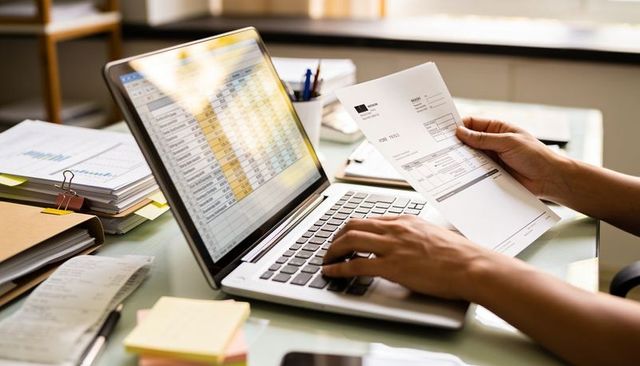 Reviewing invoices on laptop with spreadsheet and paperwork on sunlit office desk
