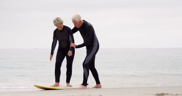 Senior couple wearing wet suits practicing surfing on beach. Man assisting woman to stand on surfboard. Ocean waves and sand in background. Ideal for concepts related to active seniors, fitness, travel, or retirement activities.