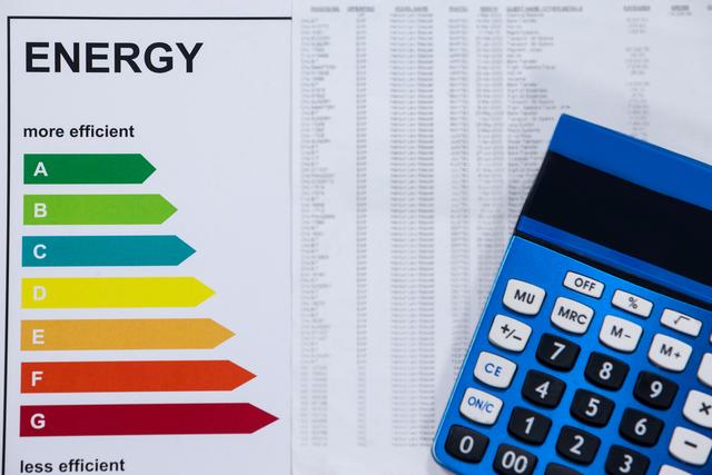 Energy efficiency rating chart next to a calculator on a wooden table. Useful for illustrating concepts related to energy consumption, cost calculation, financial planning, and sustainability. Ideal for articles, blogs, and presentations on energy saving, home improvement, and eco-friendly practices.