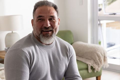 Smiling mature man sitting in cozy living room wearing light gray ribbed sweater in natural light