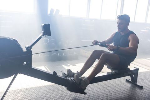 Muscular asian man rowing on indoor rower in sunlit gym demonstrating power strength endurance