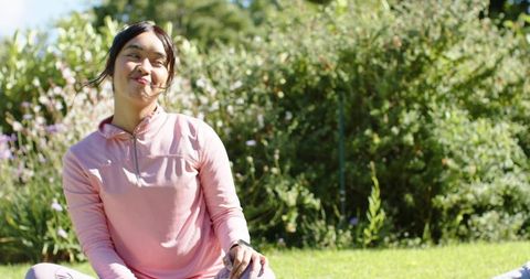 Smiling asian woman sitting cross-legged on lawn in pink pullover relaxing in sunny garden