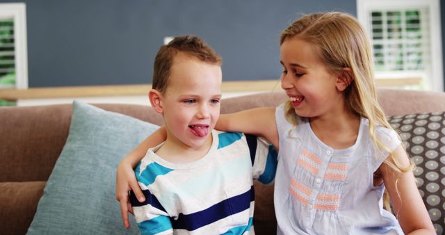 Two young siblings sitting on a sofa in a cozy living room, sharing a moment of playful fun. The brother playfully sticks out his tongue, making the sister laugh heartily. This image can be used for promoting family activities, childcare services, and home-related content highlighting joyful and light-hearted family interactions.