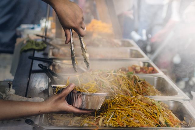 A street vendor is serving hot, steaming noodles into a takeaway container at an outdoor food market. This photo captures the essence of street food culture, showcasing a variety of dishes in serving trays. Ideal for use in articles, blogs, or advertisements related to street food, culinary experiences, food festivals, or diverse dining options.