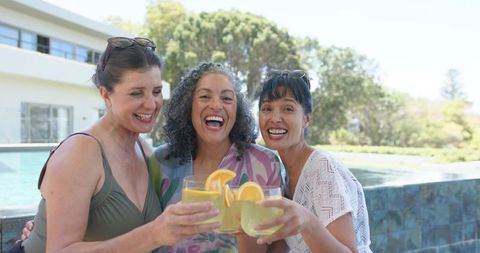 Laughing friends toasting citrus cocktails by poolside, summer resort togetherness