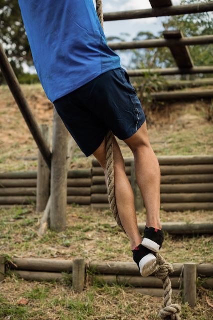 Man climbing rope during an outdoor obstacle course in a boot camp. Ideal for use in fitness, training, and adventure-related content. Can be used to illustrate themes of physical challenge, strength, and determination.