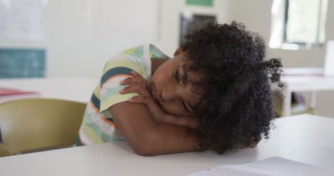 African boy resting head on folded arms at classroom desk, tired student taking break
