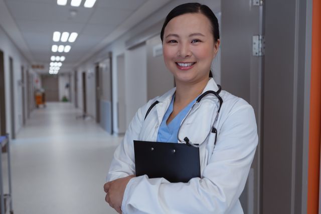 Front view of happy female doctor standing with clipboard in corridor at hospital