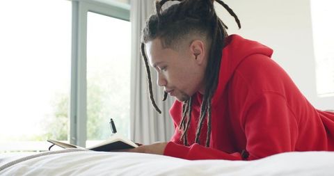 Mid-adult man with dreadlocks writing in journal on bed, red hoodie, calm morning routine