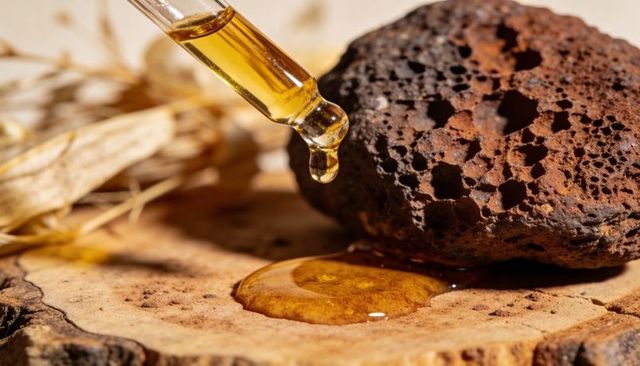 Dispensing golden facial oil droplet onto wooden slice with pumice stone and dried grass