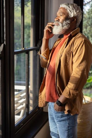 Senior man talking on smartphone by sunlit window wearing tan overshirt, jeans and smartwatch