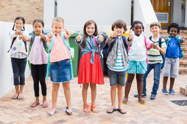 Group of cheerful kids with backpacks showing thumbs up while standing outside school building. Ideal for advertising, educational materials, back to school campaigns, and promoting childhood camaraderie.