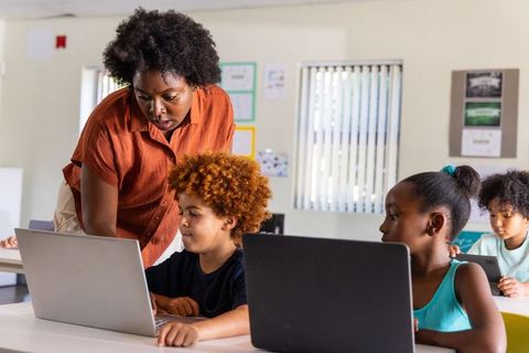 African American Teacher Guiding Elementary Kids Using Laptops in Classroom Tech Lesson
