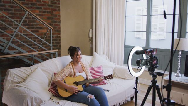 Caucasian woman sitting on sofa in cozy living room, playing guitar, recording tutorial for viewers. Room features modern decor with brick wall and large window. Picture ideal for articles or websites related to music lessons, online education, blogging tips, content creation, social media influencers, or home activities during lockdown.