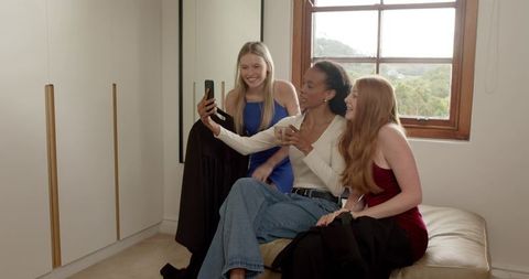 Diverse friends trying on dresses in dressing room taking selfie and laughing together