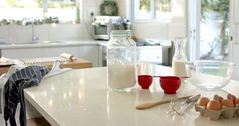 Sunlit kitchen countertop with glass flour jar, measuring cups, eggs for home baking