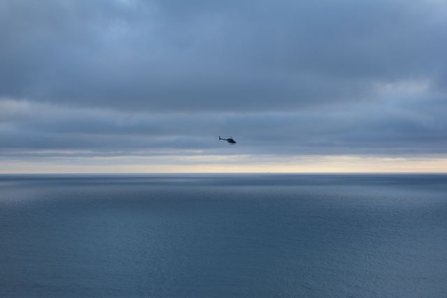 Helicopter flying low over calm ocean during dusk with a muted sky and horizon line. Ideal for travel and adventure themes, marketing materials for airlines or tour operators, and calming backgrounds.