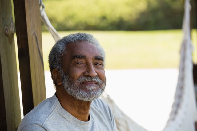 Senior man enjoying a peaceful moment outdoors, sitting near a hammock. Ideal for use in advertisements or articles related to retirement, senior lifestyle, leisure activities, and outdoor relaxation.