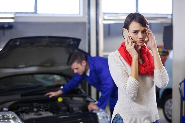 Woman standing in auto repair shop, talking on mobile phone with concerned expression while mechanic in background examines car. Useful for illustrating car trouble, customer service, vehicle maintenance, and auto repair scenarios.