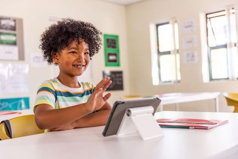 Smiling african american child waving during virtual lesson on tablet in modern classroom