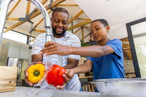 African american father teaching son washing bell peppers together in modern kitchen