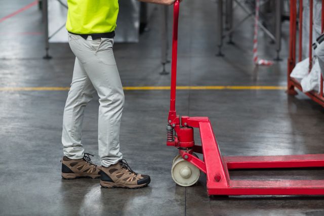 Low section of a factory worker pulling a red trolley in an industrial warehouse. The worker is wearing safety gear including work boots and light-colored pants. Ideal for use in articles or advertisements related to industrial work, warehouse logistics, safety equipment, and manual labor.