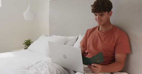 Young man sitting on bed using laptop holding passport planning trip in modern bedroom