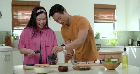 Asian couple grinding herbs and garlic with mortar and pestle in sunlit modern kitchen