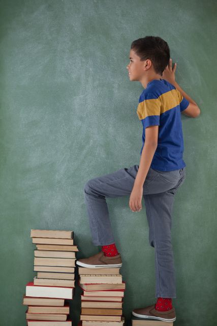 Schoolboy climbing steps of books stack against chalkboard