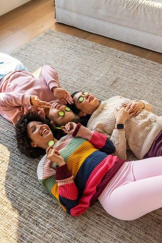 Multicultural women lying and relaxing with cucumber eye masks on woven rug in cozy home spa