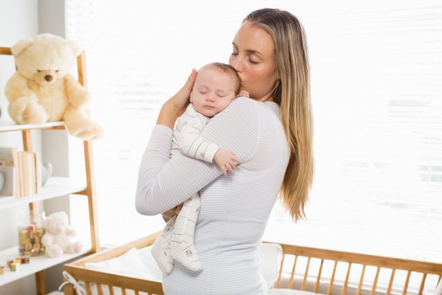 Mother holding and kissing her baby boy in a cozy home environment, showcasing love and bonding. Ideal for use in parenting blogs, family-oriented advertisements, and articles about motherhood and childcare.