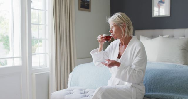 Senior woman enjoying cup of tea in cozy bedroom, dressed in white robe, sitting on bed. Bright window light creating a serene atmosphere. Ideal for concepts of relaxation, morning routine, domestic life, and tranquility. Perfect for wellness, healthcare, and lifestyle content.