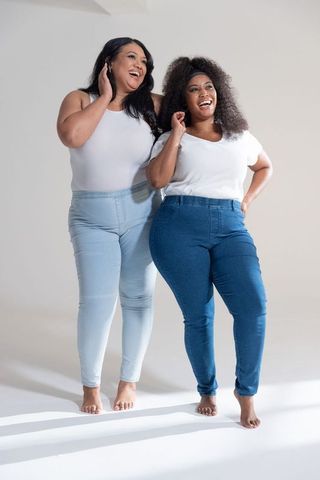 Laughing curvy friends posing in white tops and denim jeans barefoot, joyful body positive duo