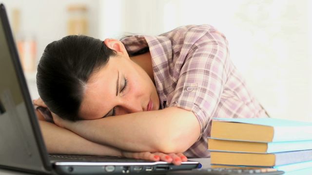 Young woman taking nap during intense study session, with head resting on table next to laptop and stack of books. Useful for articles about study tips, work-life balance, dealing with fatigue, or productivity. Can also illustrate concepts of remote work comforts.