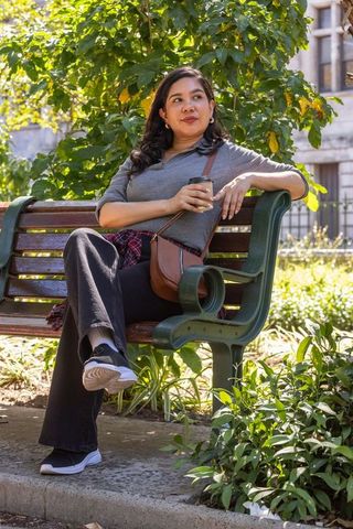Woman sitting on park bench holding coffee, gazing thoughtfully in sunlit urban garden